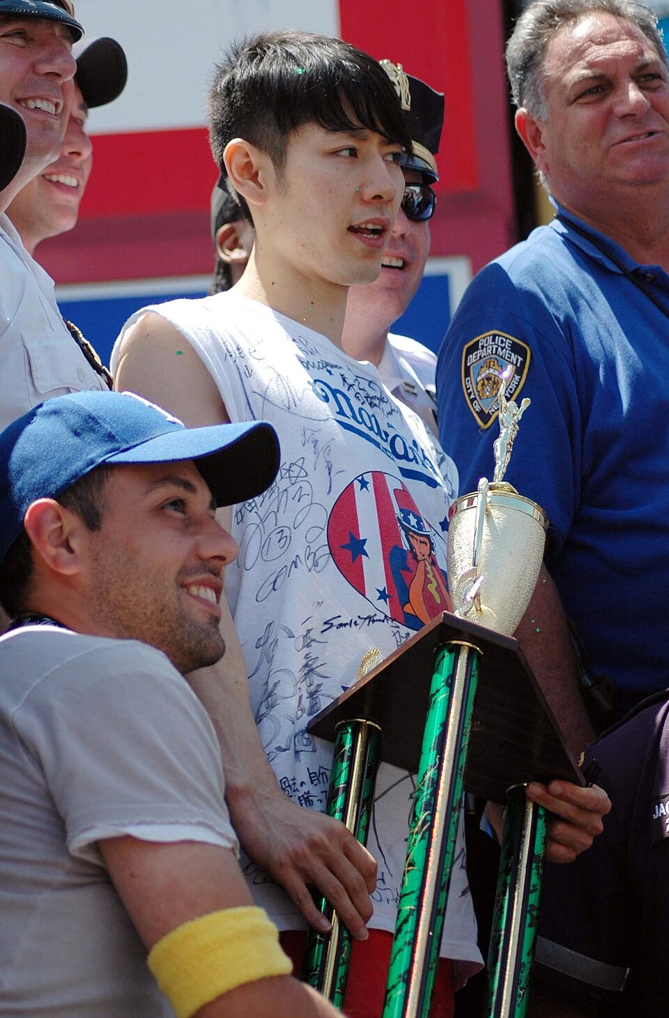 Takeru Kobayashi standing among officials and holding a trophy at a Nathan's Hot Dog Eating Contest event
